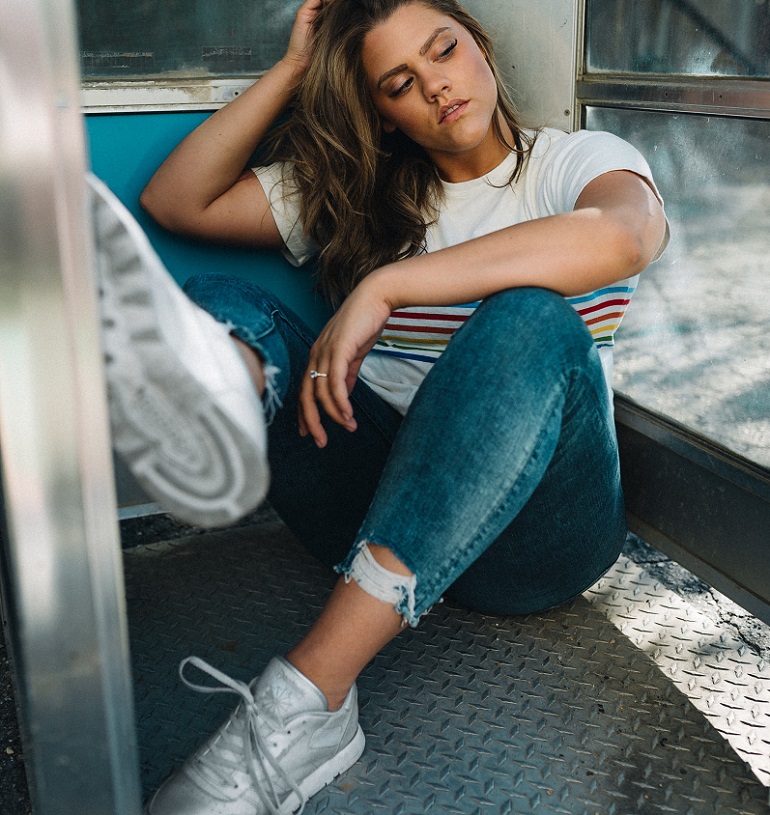 picture of  a woman in shirt, jeans and slip on sneakers sitting on a train seat 